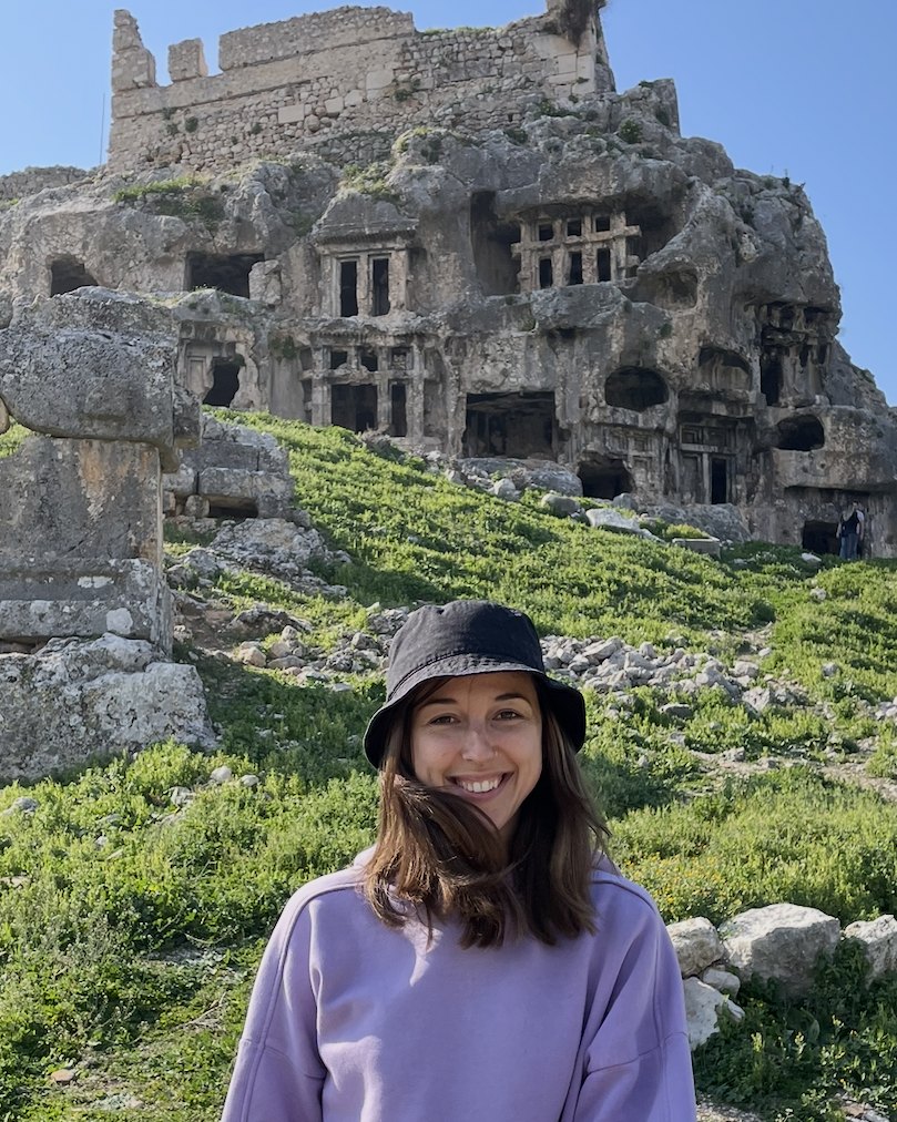 Dasha Canfield in a bucket hat, smiling, before ancient rock-cut tombs on a green hillside